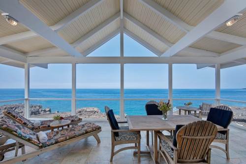 Cozy seaside patio with lounge chairs and a dining table, overlooking calm blue waters and a clear sky.