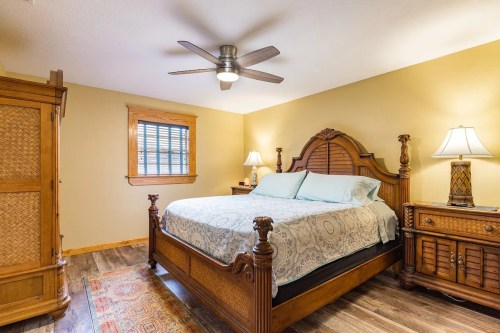 Cozy bedroom featuring a wooden bed, nightstands, lamps, and a window with blinds. Warm yellow walls and wooden flooring.
