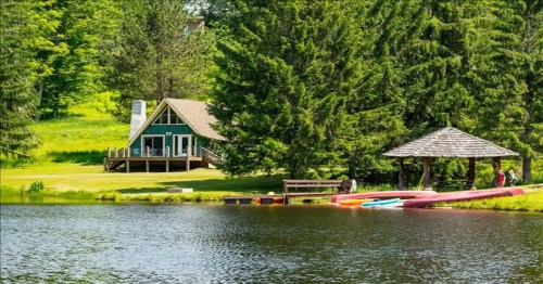 A serene lakeside scene featuring a green cabin, a gazebo, and colorful kayaks by the water, surrounded by trees.
