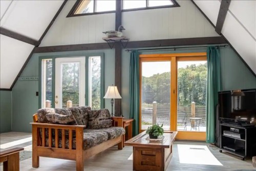Cozy A-frame living room with wooden furniture, large windows, and a view of nature outside.