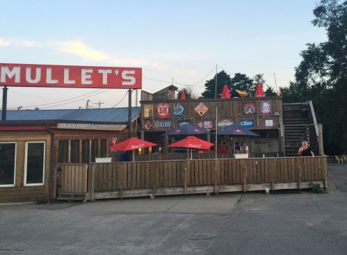 A rustic bar named "Mullet's" with outdoor seating and umbrellas, set against a twilight sky.