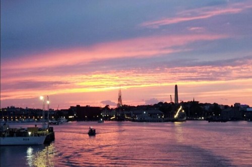 A vibrant sunset over a calm harbor, with colorful clouds reflecting on the water and silhouettes of buildings in the background.