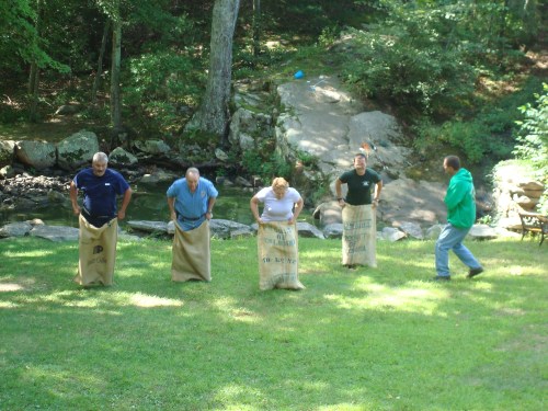 Four people in sack race bags compete on a grassy area near a stream, with one person watching from the side.
