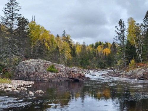 A serene river flows through a forest with autumn-colored trees under a cloudy sky.
