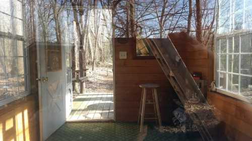 A rustic cabin interior with wooden walls, a stool, and an open door leading to a forested area outside.