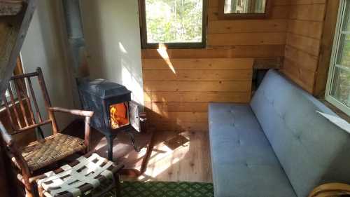 Cozy interior of a cabin with a gray sofa, wooden chair, and a small stove, illuminated by natural light.