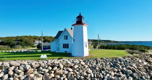 A white lighthouse stands on a grassy shore, surrounded by rocks, with a clear blue sky and ocean in the background.