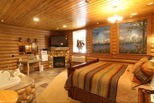 Cozy log cabin bedroom with a fireplace, large bed, and scenic mountain artwork on the walls.