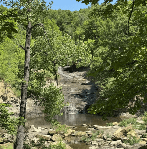 A serene waterfall cascades down rocky terrain, surrounded by lush green trees and a calm pool below.