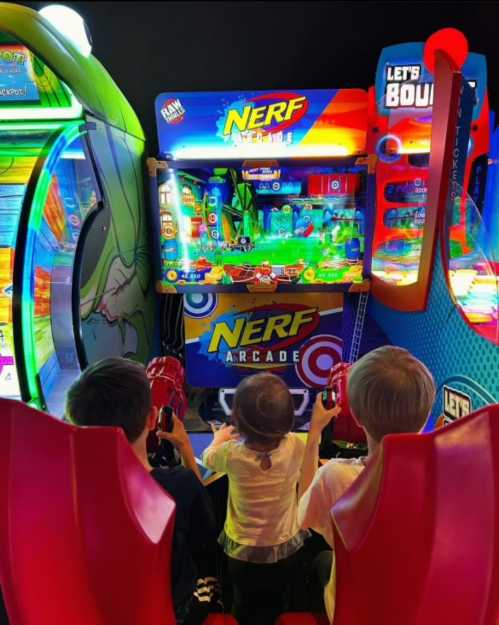 Three children play a colorful Nerf arcade game in a vibrant arcade setting.