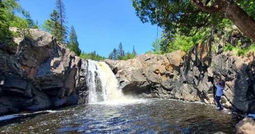 A scenic waterfall cascades over rocky cliffs into a serene pool, surrounded by lush greenery and tall trees.
