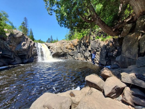 A scenic waterfall cascades into a dark pool, surrounded by rocky cliffs and lush greenery under a clear blue sky.