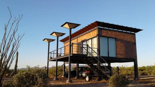 A modern, elevated house with large windows and a deck, surrounded by desert vegetation and clear blue skies.