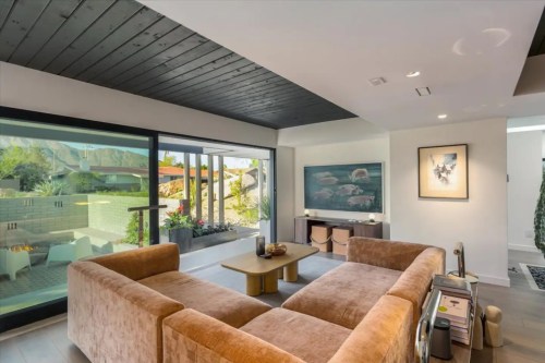 Modern living room with a brown sofa, large windows, and a view of outdoor greenery. Minimalist decor and artwork.