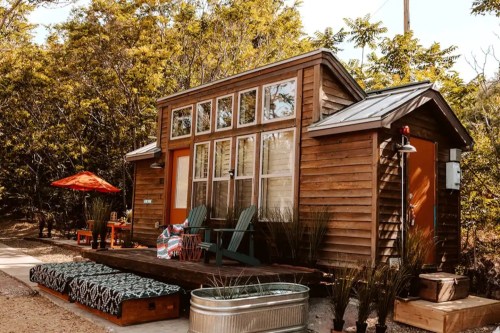 A cozy wooden tiny house with large windows, surrounded by greenery, featuring a deck and outdoor seating area.
