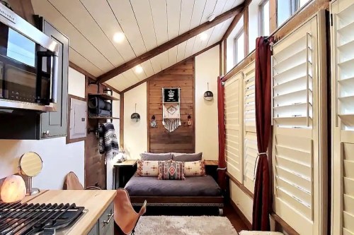 Cozy tiny home interior featuring a sofa, kitchen area, and large windows with wooden accents and warm decor.