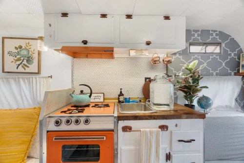 Cozy vintage kitchen in a camper, featuring an orange stove, white cabinets, and decorative plants.