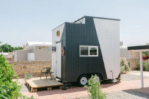 A modern tiny house on wheels with a black and white exterior, parked on a stone patio surrounded by greenery.