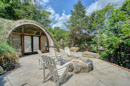 A modern, curved structure with a grassy roof, surrounded by trees and a stone fire pit with white chairs.