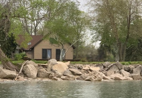 A lakeside cabin surrounded by trees and large rocks, with a calm water view in the foreground.
