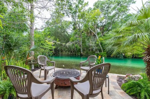 A serene outdoor space with chairs around a fire pit, overlooking a lush green river surrounded by trees.