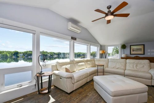 Bright living room with a white sectional sofa, large windows overlooking a serene lake, and a ceiling fan.