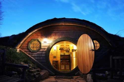 A cozy hobbit-style home with a round door, illuminated interior, and a twilight sky in the background.
