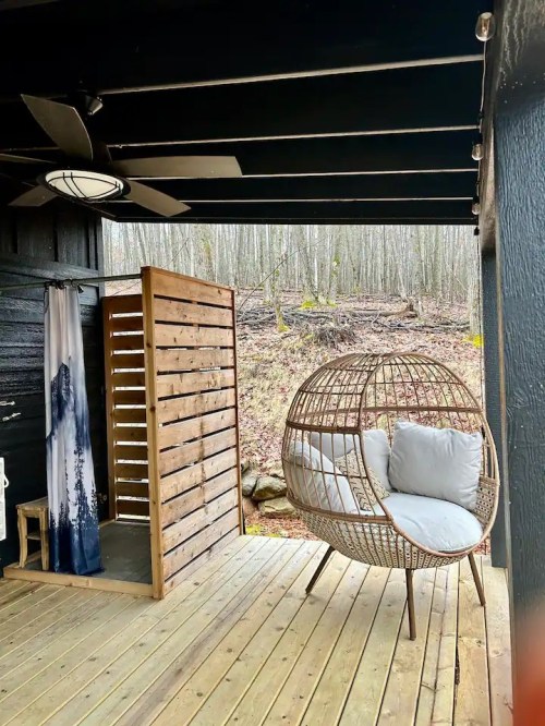 A cozy outdoor space featuring a wooden shower, a round wicker chair, and a serene forest backdrop.