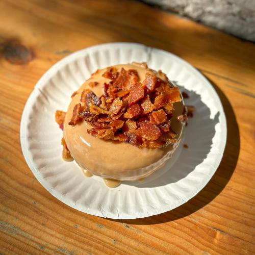 A glazed donut topped with crispy bacon bits on a paper plate, resting on a wooden surface.