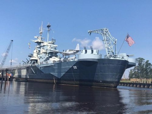 A large naval ship, the USS North Carolina, docked by the water with an American flag flying. Clear blue sky in the background.