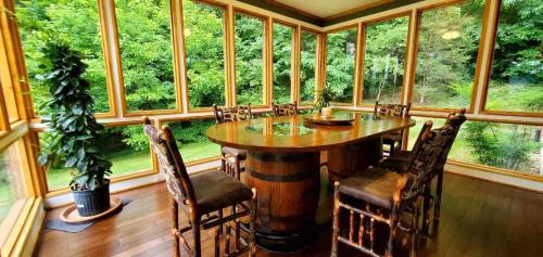 A bright sunroom with large windows, featuring a wooden table and chairs, surrounded by greenery outside.