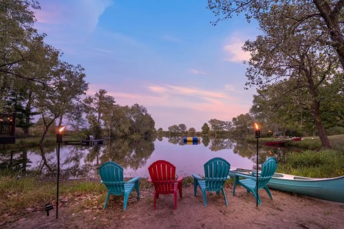 Four colorful chairs by a serene lake at sunset, with torches and a canoe nearby, surrounded by trees.
