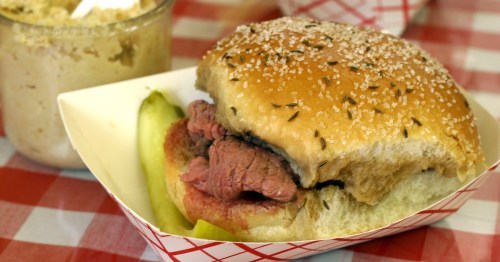 A close-up of a sandwich with sliced meat on a bun, garnished with herbs, served with a pickle on a checkered tablecloth.
