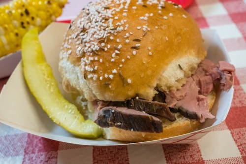 A close-up of a sandwich with sliced meat on a sesame seed bun, accompanied by a pickle slice on a checkered tablecloth.