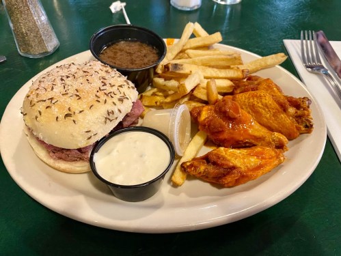 A plate with a sandwich, fries, chicken wings, and two dipping sauces on a green table.