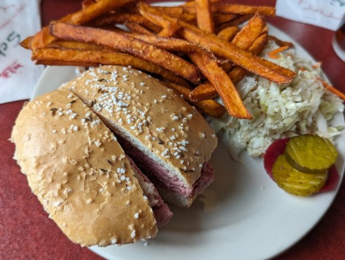 A plate featuring a sesame seed bun sandwich with meat, sweet potato fries, coleslaw, and pickles.