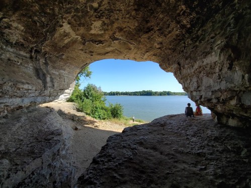 A view from inside a cave opening, overlooking a river and lush greenery, with two people sitting nearby.