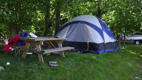 A blue and gray tent set up on grassy ground near a picnic table and camping chairs under leafy trees.