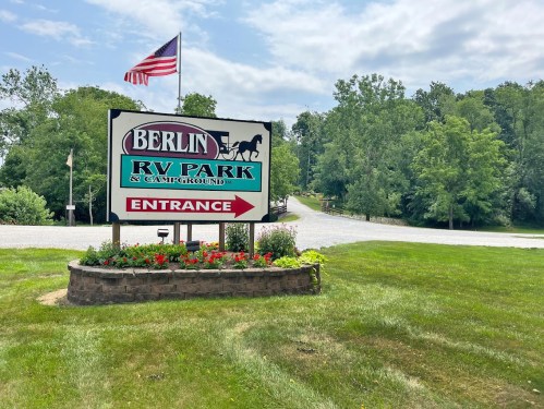 Sign for Berlin RV Park & Campground with an American flag, surrounded by greenery and flowers, indicating the entrance.
