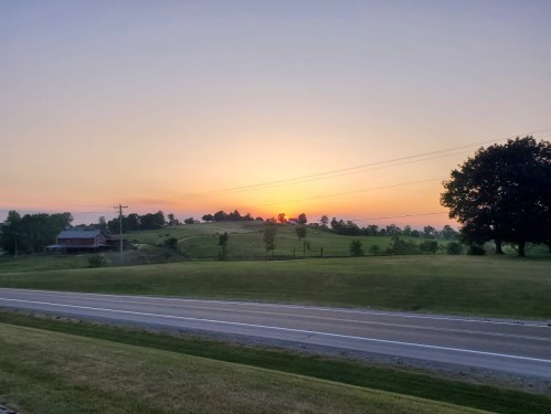 A serene landscape at sunset, featuring rolling hills, a distant barn, and a quiet road lined with trees.
