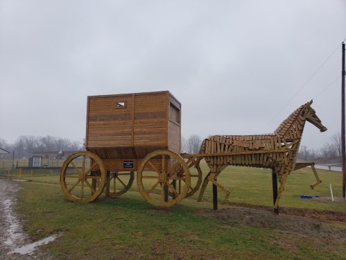 A wooden sculpture of a horse pulling a cart, set in a grassy area under a cloudy sky.