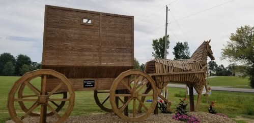 A large wooden sculpture of a horse and wagon, set in a grassy area with trees in the background.