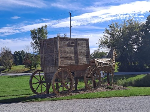 A large wooden sculpture of a horse-drawn wagon on a grassy area, with trees and a blue sky in the background.