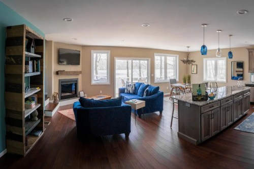 A cozy living room with blue furniture, a fireplace, and large windows, featuring a kitchen area in the background.