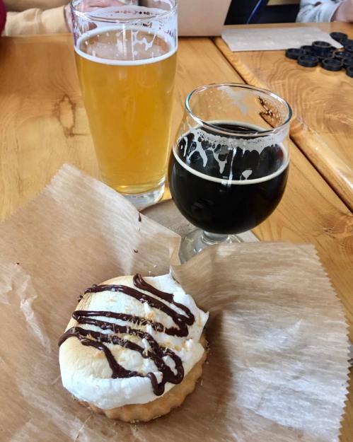 A cookie topped with whipped cream and chocolate drizzle, accompanied by two glasses of beer on a wooden table.