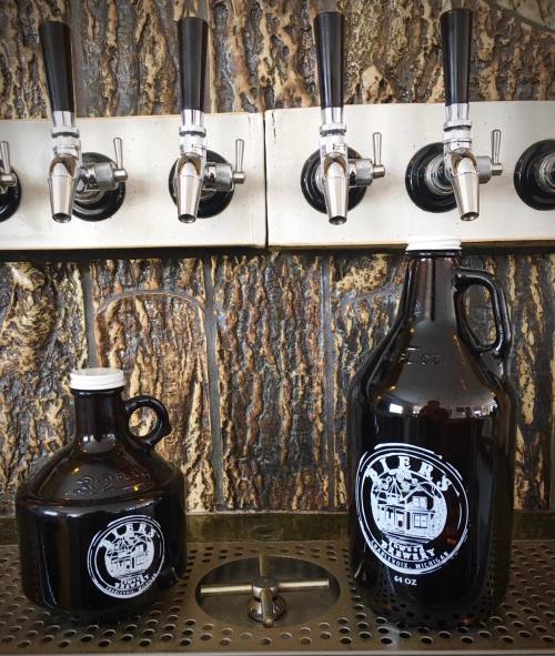 Two dark glass growlers sit on a counter beneath beer taps, showcasing a rustic brewery setting.