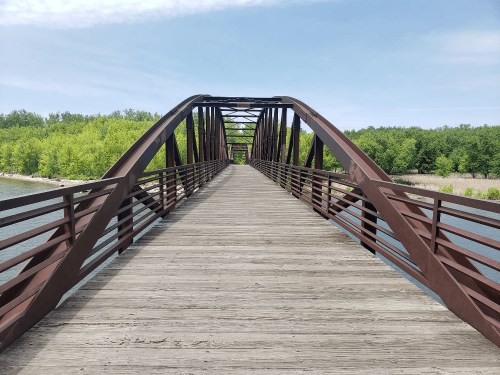 A wooden bridge with a metal arch design spans over a calm body of water, surrounded by lush green trees.