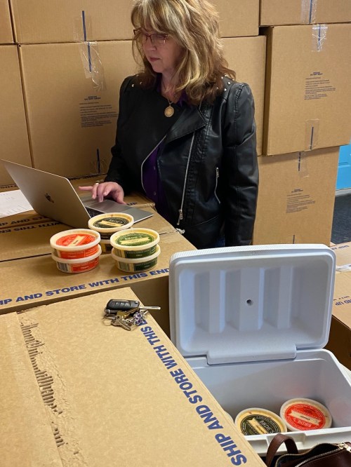 A woman in a leather jacket works on a laptop beside stacked boxes and containers of food.