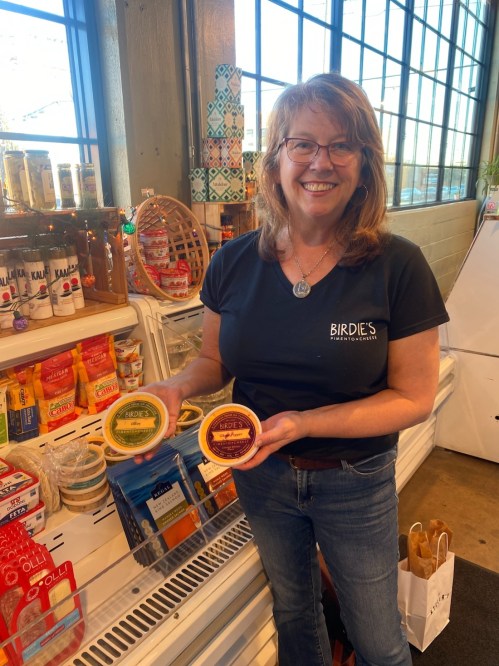 A smiling woman holds two containers of hummus in a market, surrounded by various food products on display.
