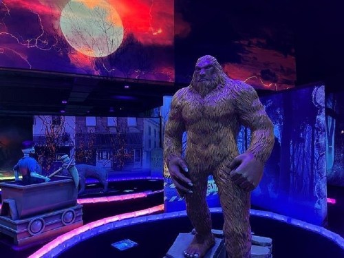 A large, furry Bigfoot statue stands in a colorful, illuminated exhibit with a dramatic moonlit backdrop.